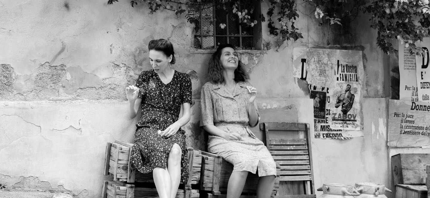 Na zdjęciu dwie kobiety siedzące na skrzyniach pod scianą.