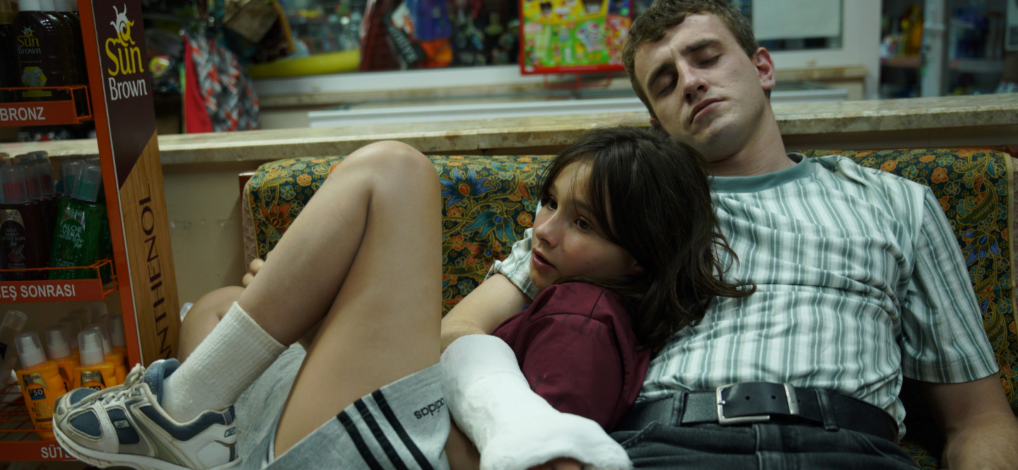 Na zdjęciu młody mężczyzna siedzący na kanapie. Ma zamknięte oczy. Ręką w gipsie obejmuje dziewczynkę opierającą się o niego plecami.