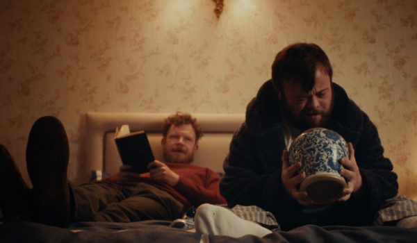 Na zdjęciu mężczyzna siedzący na łóżku zagląda do ceramicznej wazy. Z tyłu leży drugi mężczyzna i mu się przygląda.