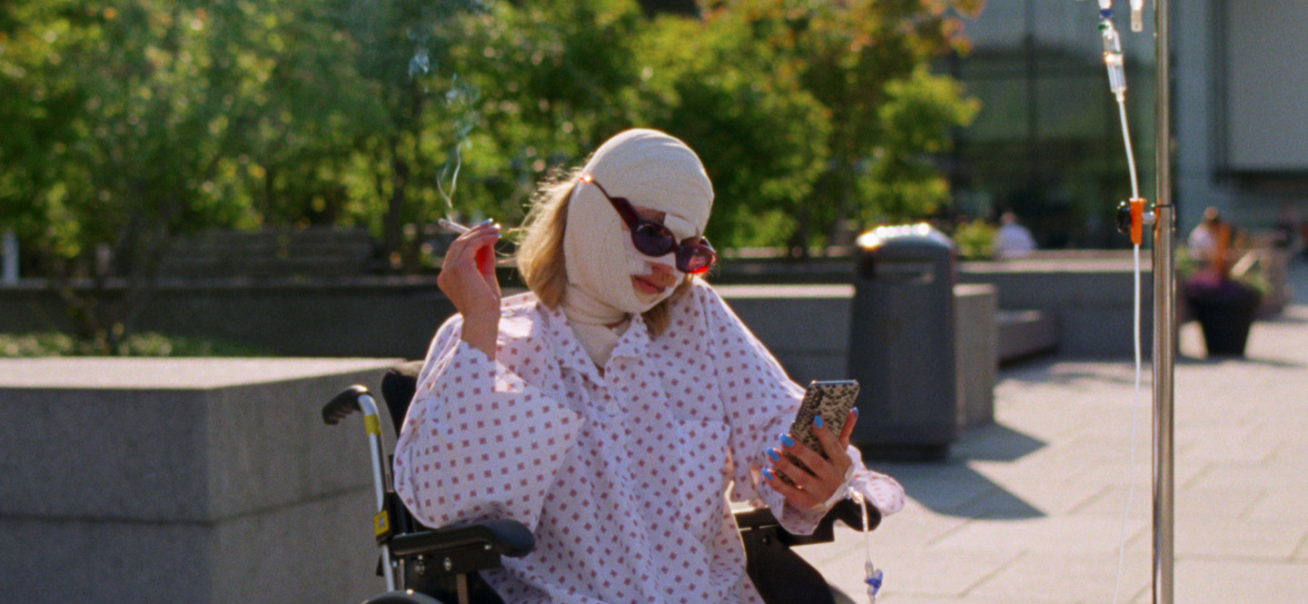 Na zdjęciu kobieta z zabandażowaną głową siedzi na wózku inwalidzkim pali papierosa i przegląda telefon.