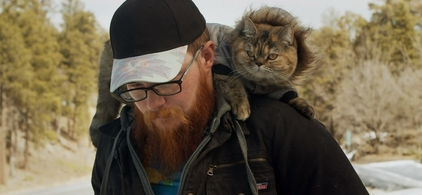 Na zdjęciu młody, brodaty mężczyzna w czapce z daszkiem. Na jego ramionach siedzi kot.