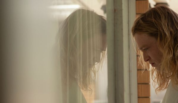 Na zdjęciu młody mężczyzna w długich blond włosach. Stoi z głową pochyloną na przeciwko okna. W szybie odbija się jego twarz.