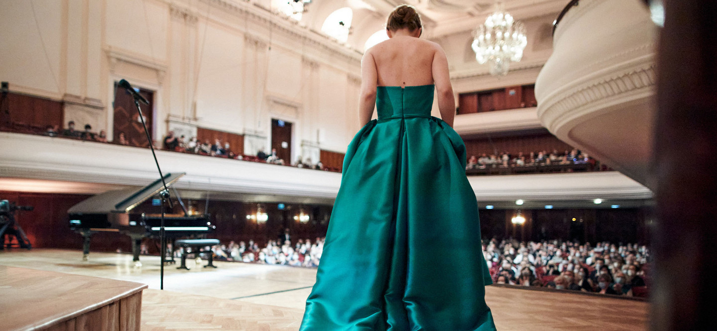 Na zdjęciu stojąca tyłem do kadru kobieta w zielonej, wieczorowej sukni kłaniająca się publiczność w sali koncertowej.