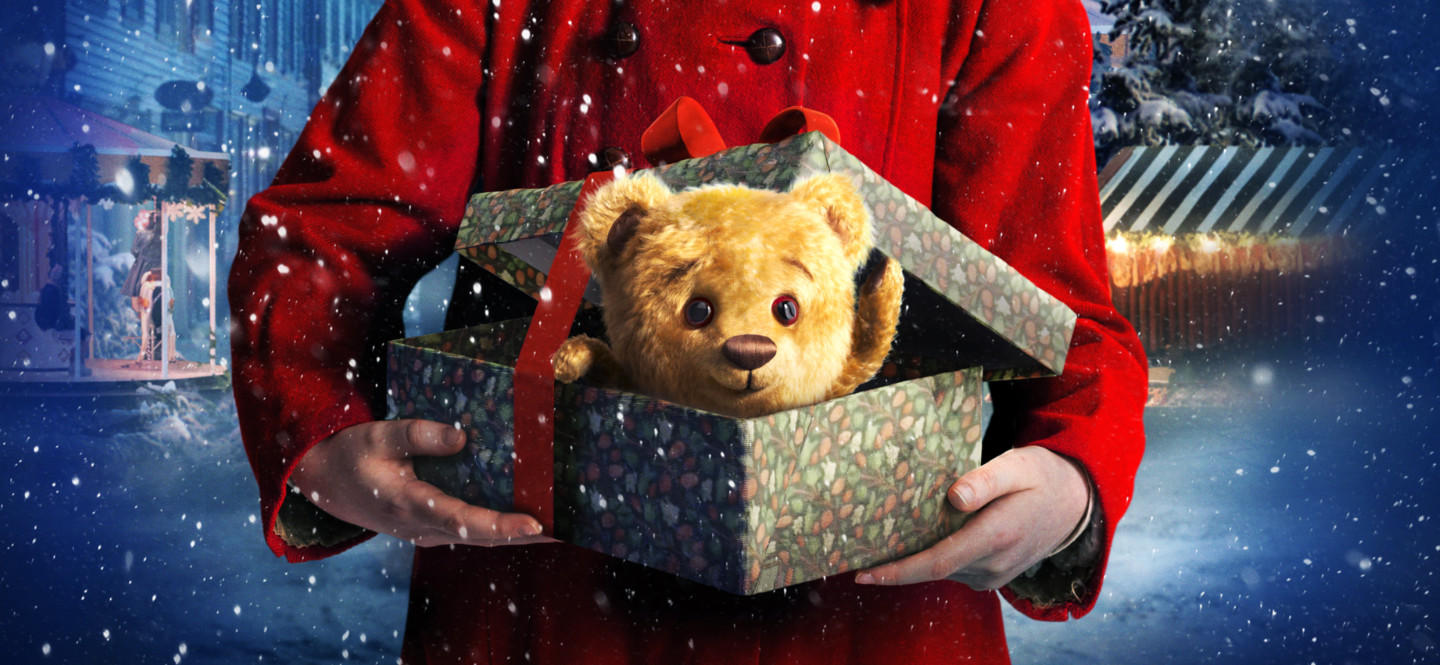 Na zdjęciu osoba w czerwonym płaszczu trzyma świąteczne pudełko. Z pudełka wychyla się pluszowy miś.